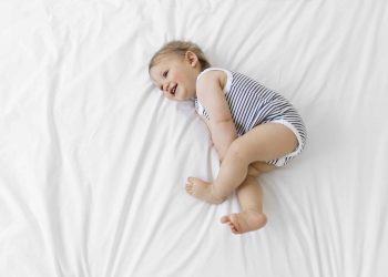 High Angle View Of Baby Boy Wearing Striped Onesie Lying On Bed With White Duvet.